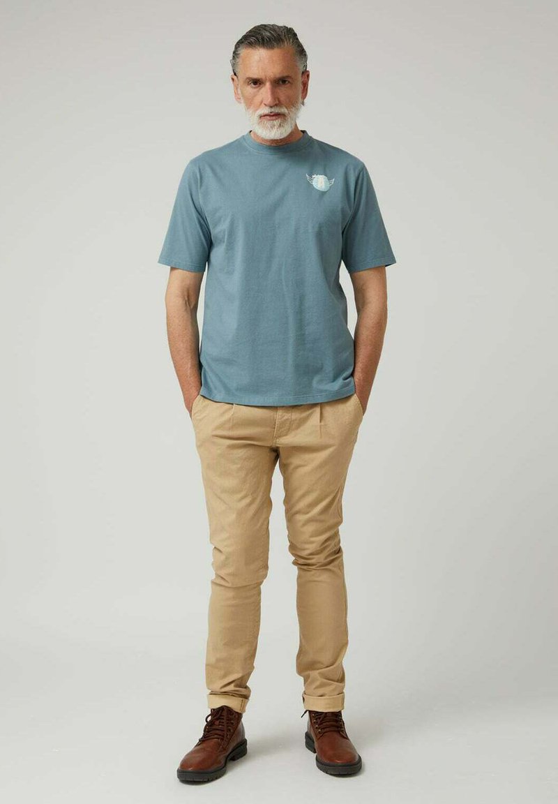 Camiseta de algodón azul claro con mangas cortas, con un pequeño estampado en el pecho. Pantalones beige de corte ajustado y botas marrones con cordones.