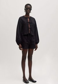 Zwart textuuroutfit met een cropped top met een strik aan de voorkant, lange mouwen en bijpassende shorts. Model draagt platte schoenen en een zonnebril.