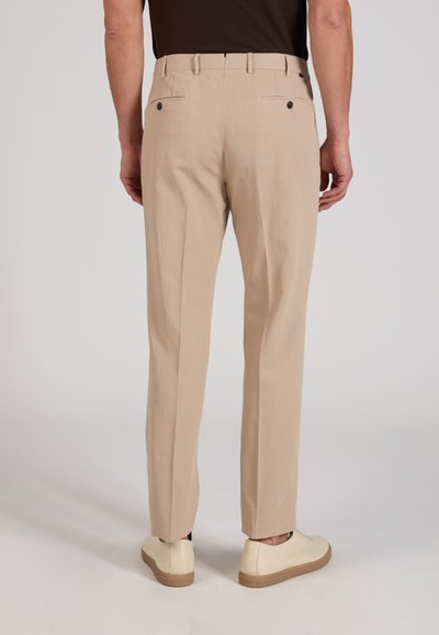 Homme portant un pantalon beige sur mesure et des baskets écru, debout devant un fond uni, vu de dos.