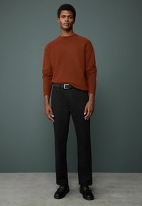 Rostfarbener Strickpullover aus strukturiertem Material, kombiniert mit schwarzen Hosen und schwarzen Schuhen mit Hardware-Akzenten. Schlichtes Design, figurbetonter Schnitt.