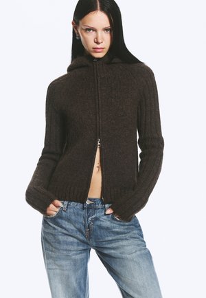 Jeune femme aux longs cheveux bruns foncés portant un pull zippé marron foncé et un jean bleu, mains dans les poches, avec une expression neutre.