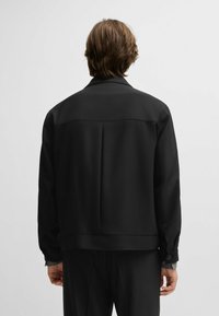 Giacca nera con una texture liscia, dotata di colletto a punta, una piega sul retro e polsini abbottonati. Design semplice e strutturato.