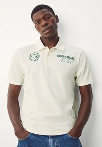 Cremfarbener Poloshirt mit weicher Textur. Mit grünem gesticktem "Snoopy Social Club" Logo und Lorbeerdesign auf der linken Brust. Kurzärmelig.
