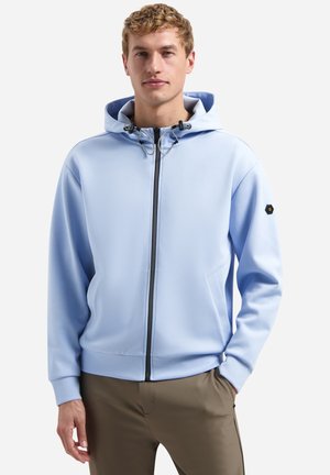 Man draagt een lichtblauwe hoodie met rits en trekkoorden en beige broek, staat met één hand in de zak tegen een effen achtergrond.