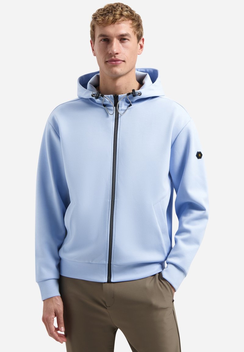 Man draagt een lichtblauwe hoodie met rits en trekkoorden en beige broek, staat met één hand in de zak tegen een effen achtergrond.