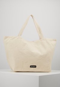 Beige canvas tote bag with two handles, rectangular base, and small black label reading "KARL LAGERFELD" on front, placed on white surface.