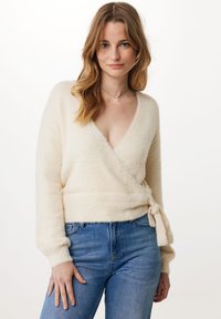 Cremfarbener Wickelpullover aus weichem, flauschigem Material. Mit tiefem V-Ausschnitt, langen Ärmeln und einer gebundenen Taillendetail. Kombiniert mit blauen Jeans.