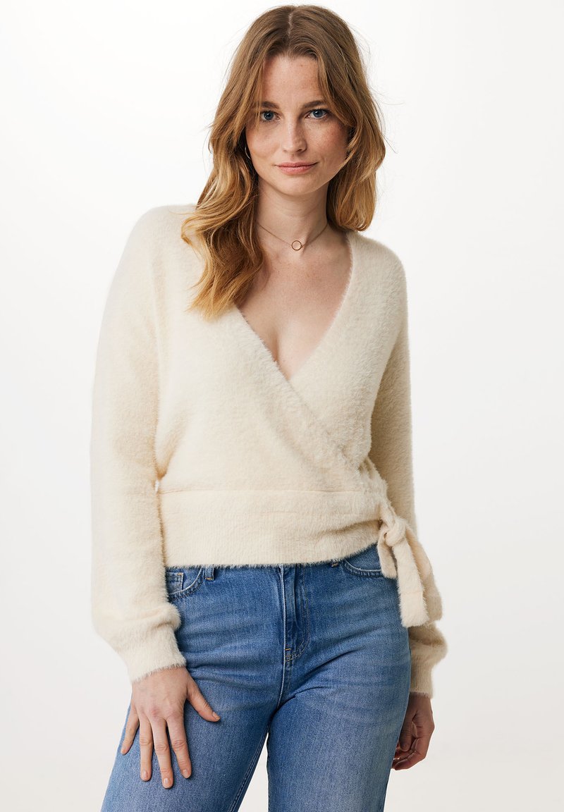 Cremfarbener Wickelpullover aus weichem, flauschigem Material. Mit tiefem V-Ausschnitt, langen Ärmeln und einer gebundenen Taillendetail. Kombiniert mit blauen Jeans.