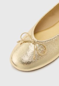 Χρυσό μεταλλικό μπαλαρίνα παπούτσι με στρογγυλεμένη μύτη και δεμένη φιόγκο στην κορυφή, με απαλό μπεζ εσωτερικό επένδυση.