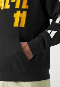 Sweat à capuche noir avec un lettrage jaune vif, poche kangourou et accents blancs sur les manches. Texture douce, coupe décontractée et poignets côtelés.