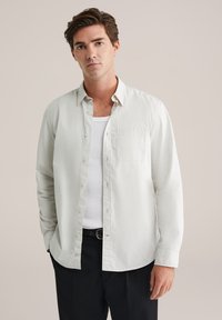 Šviesios spalvos medvilninis marškinėlių mygtukais viršus su krūtinės kišene, laisvu siluetu ir ilgomis rankovėmis, dėvimas virš baltos marškinėlių.