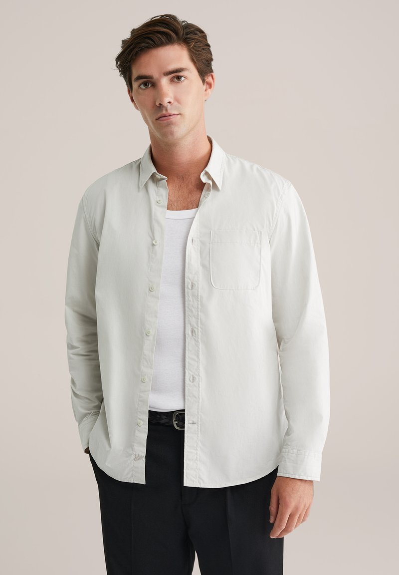 Šviesios spalvos medvilninis marškinėlių mygtukais viršus su krūtinės kišene, laisvu siluetu ir ilgomis rankovėmis, dėvimas virš baltos marškinėlių.