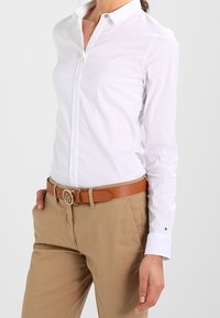 Weißes langärmliges Hemd mit Kragen, kombiniert mit beigen Hosen und einem braunen, strukturierten Gürtel mit einer glänzenden Schnalle.