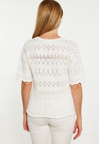 Top bianco lavorato a maglia con maniche corte, presenta intricate fantasie a diamante e orizzontali, orlo ondulato e una finitura testurizzata.