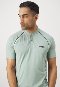 Koszulka polo z krótkim rękawem, z kołnierzykiem w jasnym miętowym kolorze, z czarnymi i białymi akcentami. Logo "BOSS" na piersi. Gładka tekstura.