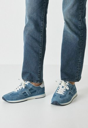 Blaue Wildleder-Sneaker mit weißen Schnürsenkeln, grauem Akzent und strukturierten Streifen. Kombiniert mit dunklen Denim-Jeans, die einen lässigen Stil zeigen.