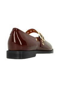 Chaussure en cuir marron avec une finition brillante, bout arrondi et un petit talon noir. Elle possède une boucle sur le côté et des détails perforés le long de la sangle.