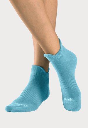 Füße mit hellblauen Knöchelsocken mit dem "Bench"-Logo, ein Fuß angehoben und über den anderen geschlagen vor einfarbigem Hintergrund.