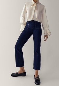 Vit satinblus med en slips och rynkade manschetter, kombinerad med mörkblå högmidjade jeans och svarta läderlovers.