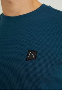 Blå texturerad tyg sweatshirt med rund halsringning, försedd med en svart fyrkantig patch med vit detaljer på bröstet.
