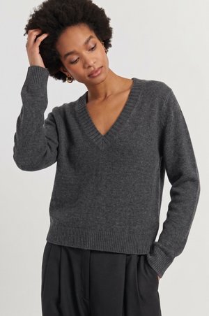 Vrouw met kort krullend haar, die een donkergrijze V-hals trui en zwarte broek draagt, met één hand haar haar aanrakend.