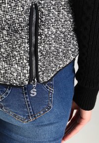 Sort og hvid tekstureret vest med lynlås, parret med blå jeans med baglomme-syning.