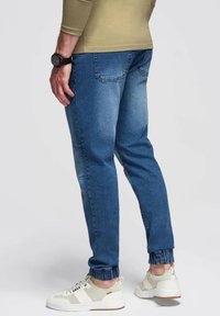 Joggers de mezclilla en azul medio con pierna ajustada, tobillo cuffado y bolsillos traseros. Combinados con zapatillas de color claro.