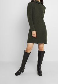 Kvinna i en mörkgrön långärmad stickad klänning och svarta knähöga klackiga läderstövlar, stående mot en enkel ljus bakgrund.