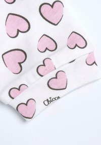 Vitt tyg med rosa hjärtan i svart kontur. Har ribbade ärmslut och en etikett som säger "Chicco." Slät textur, avslappnad design.