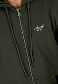 Sudadera con capucha de cremallera en verde oliva, confeccionada en un tejido suave, con una cremallera plateada y el logotipo bordado en blanco "Reell est. 1997" en el pecho.