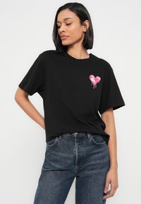 Μαύρο βαμβακερό T-shirt με στρογγυλή λαιμόκοψη, κοντά μανίκια και ένα ροζ καρδιά που στάζει γραφικό στο αριστερό μέρος του στήθους. Φοράει με ψηλόμεσο μπλε τζιν.