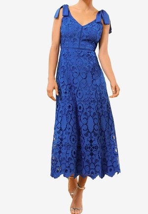 Kobieta w niebieskiej, koronkowej sukience midi bez rękawów z kokardkowymi ramiączkami i srebrnymi, otwartymi na palce butami na obcasie, stojąca na jednolitym białym tle.