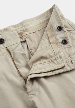 Beige katoenen broek met een rits aan de voorkant en twee zwarte knopen. Heeft een gladde textuur en een tailleband met stiksel. 