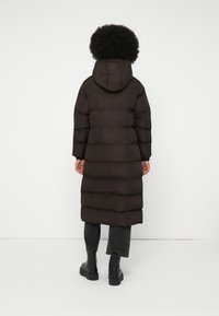 Un lungo piumino marrone scuro con cappuccio, caratterizzato da un design trapuntato e una silhouette dritta, abbinato a stivali neri alla caviglia e pantaloni grigi.