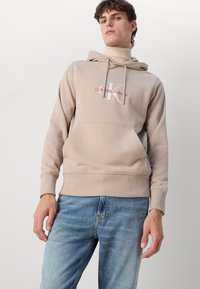 Felpa beige con cappuccio e tasca frontale, con logo "CK" bianco e testo "Calvin Klein Jeans" in rosso. Texture morbida e liscia.
