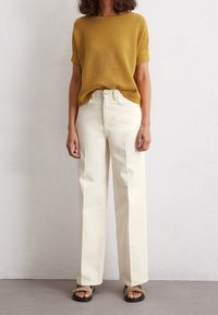 Camisa de punto texturizada en mostaza amarilla de manga corta, combinada con pantalones de pierna ancha en color crema. Sandalias negras con detalles de tiras completan el conjunto.