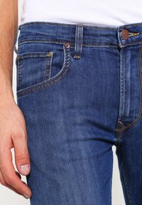 Blå denimjeans med rak passform. Har orange sömmar, en myntficka, en metallknapp och en läderloggopatch vid midjan.