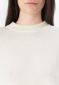 Jasnobeżowy sweter z dzianiny z prążkowanym okrągłym dekoltem. Materiał wydaje się miękki, o delikatnej fakturze i subtelnych odcieniach koloru.