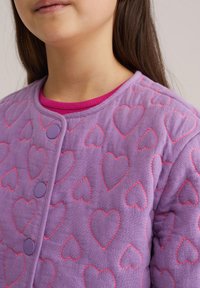 Violetti tikattu takki, jossa sydänkuvio punaisilla saumoilla, pyöreillä violeteilla napeilla ja vaaleanpunaisella resorikauluksella.
