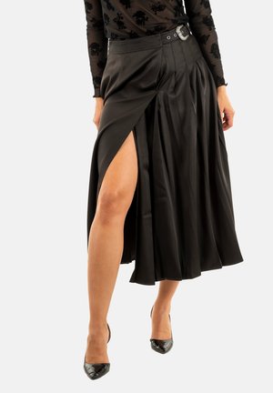Mujer que lleva una falda midi negra plisada con una gran abertura y un cinturón decorativo, combinada con una blusa negra transparente con flores y tacones de punta negros.