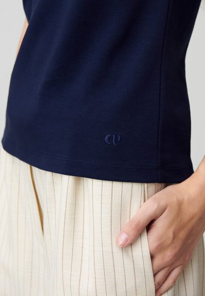 T-shirt in cotone blu navy con scollatura rotonda, caratterizzata da un logo ricamato discreto sull'orlo. Abbinata a pantaloni a righe chiare con pieghe.
