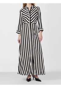 Femme portant une longue robe boutonnée à rayures noires et blanches avec des manches trois-quarts et des talons noirs à bout pointu, debout sur un sol gris.