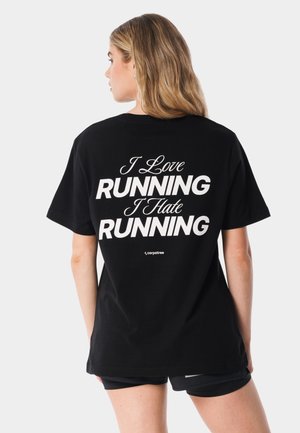 Kobieta o blond włosach ubrana w czarny t-shirt z białym napisem "I Love RUNNING I Hate RUNNING" na plecach, stojąca na jednolitym tle.