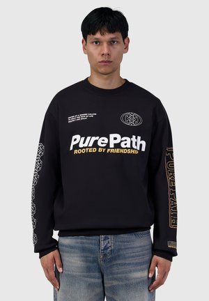 Young man wearing a black sweatshirt with white and yellow text "PurePath Rooted by Friendship" and patterned designs on sleeves, paired with blue jeans.