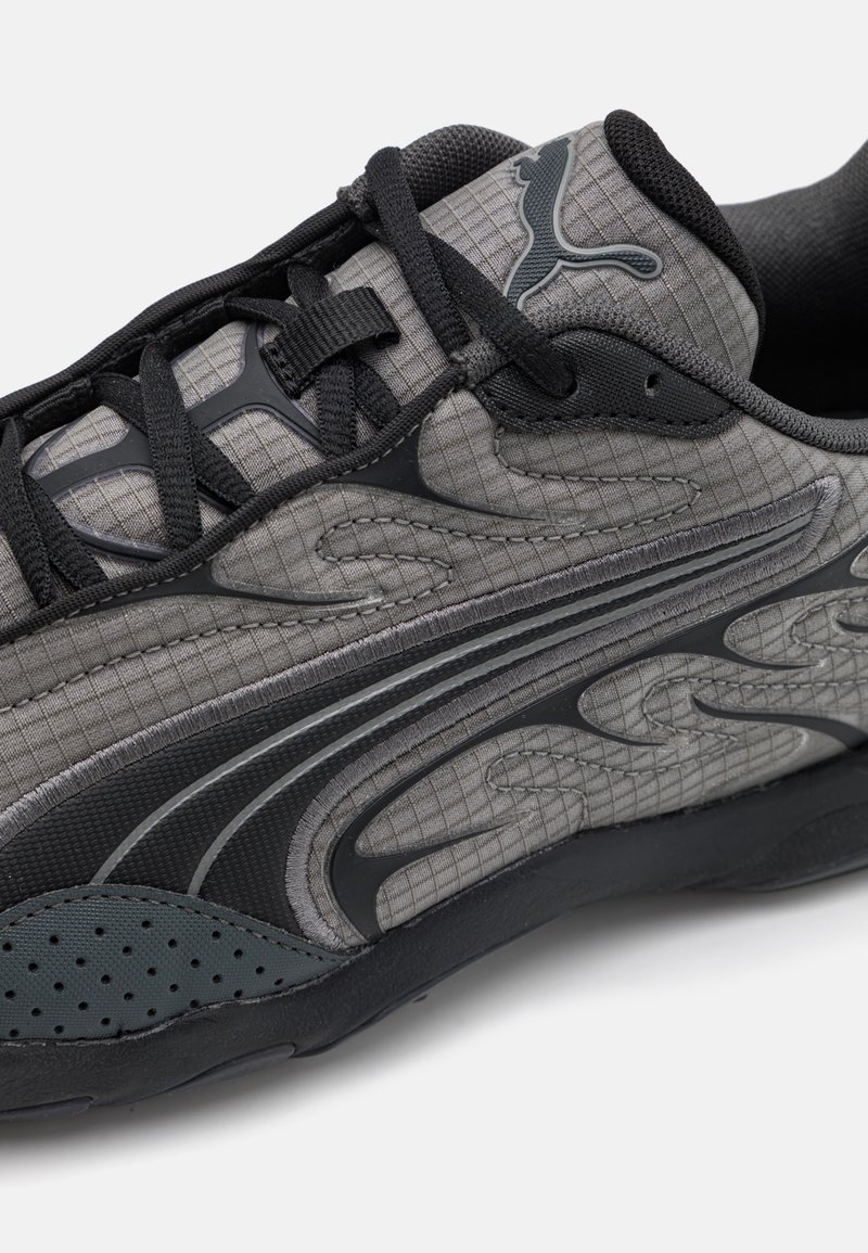 Graue Sportschuh mit strukturierten Oberflächen, schwarzen Akzenten, gepolstertem Kragen und einem gemusterten Overlay. Die Schnürsenkel sind schwarz und sicher gebunden.