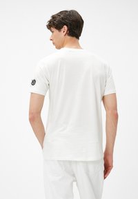Camiseta de algodón blanca con mangas cortas, cuello redondo y detalle del logo en la manga izquierda, combinada con pantalones blancos. Textura suave.