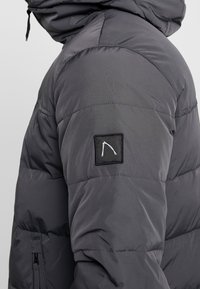 Graue Stepp-Steppjacke mit Kapuze, mit gestepptem Design, seitlichen Reißverschlusstaschen und einem schwarzen Logo-Patch auf dem linken Ärmel.