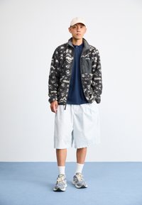 Texturiertes schwarzes und weißes Fleece-Jackett mit einer grauen Tasche, kombiniert mit einem marineblauen Polo, hellen Shorts und mehrfarbigen Sneakers.