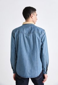 Light blue denim shirt with a contrasting beige collar, long sleeves, and a curved hem. Features buttoned cuffs and a back yoke seam design.