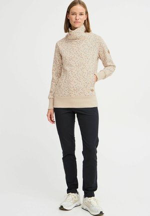 Sweatshirt beige avec un col haut et une impression florale colorée. Poignets et ourlet côtelés. Associé à un pantalon noir et des baskets blanches.